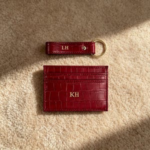 Puede incluir: Un tarjetero de cuero rojo oscuro con relieve de cocodrilo y un llavero a juego. El tarjetero presenta las iniciales KH en dorado, mientras que el llavero rectangular muestra las iniciales LH en dorado y una anilla de metal dorado.