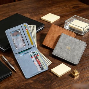 May include: A light blue bifold wallet with a clear ID window and multiple card slots containing paper currency and cards. Beside it are a brown bifold wallet and a gray bifold wallet with the gold embossed initials BK on the front cover.