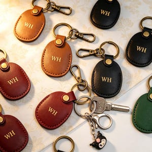 Könnte beinhalten: Eine Sammlung ovaler Schlüsselanhänger aus Leder in Braun, Schwarz, Rot und Grün, jeweils mit den geprägten goldenen Buchstaben WH. Die Anhänger verfügen über bronzefarbene Metallringe und Karabinerverschlüsse, wobei ein Schlüssel befestigt ist.