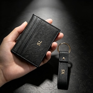 May include: A black textured card holder with the gold embossed letters SL and a matching black textured keychain with the gold embossed letters XS, featuring a gold metal ring and a small gold rivet detail on the strap.
