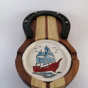 May include: A wooden ashtray featuring a central circular ceramic insert with a hand-painted illustration of a red and blue sailing ship. The wooden base is accented with a black metal horseshoe attached to the top, creating a decorative frame.