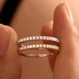 May include: Two thin metal rings stacked vertically, each featuring a continuous row of small, clear, round-cut stones set along the outer band. The top ring appears silver-toned, while the bottom ring has a warm, gold-toned metallic finish.