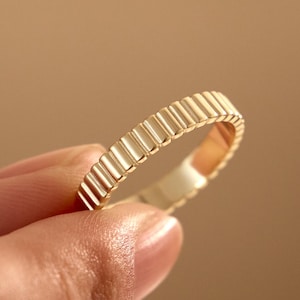 May include: A gold ring featuring a continuous, repeating vertical ribbed or fluted texture around the entire outer band. The metal has a polished, reflective finish that catches the light, creating a series of small, uniform highlights and shadows.