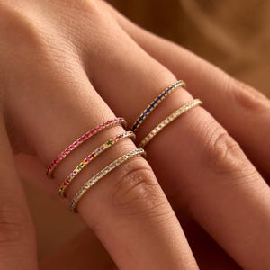 Könnte beinhalten: Ein Set aus fünf dünnen Metallringen an den Fingern. Drei Ringe sind an einem Finger gestapelt und mit rosa, bunten und klaren Steinen besetzt. Zwei weitere Ringe befinden sich an einem anderen Finger, einer mit blauen und einer mit klaren Steinen.