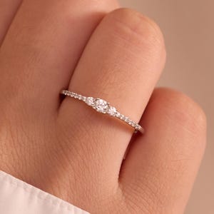 Può includere: Un sottile anello color argento con una pietra centrale rotonda e trasparente affiancata da due pietre più piccole, con minuscole pietre trasparenti incastonate lungo la fascia sottile, per un aspetto delicato e raffinato al dito.