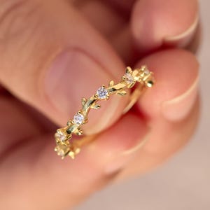 May include: A gold ring featuring a delicate vine-like band design with small, clear, round-cut stones set at intervals along the metal surface. The band has a textured, organic appearance resembling intertwined branches or leaves in a polished finish.