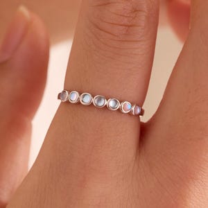 Può includere: Un sottile anello in metallo caratterizzato da una serie di piccole pietre rotonde e iridescenti incastonate in fila lungo la fascia. Le pietre mostrano sottili sfumature blu e rosa e la fascia ha una finitura metallica lucida, indossato al dito.