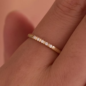 Puede incluir: Un anillo de banda fina de oro con una serie de pequeñas piedras redondas y transparentes engastadas en fila en la parte superior. La superficie metálica del anillo tiene un acabado texturizado y acanalado que recorre toda la banda con detalle.