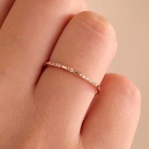 Op de afbeelding: Een dunne ring van roségoud met een delicaat afwisselend patroon van kleine ronde doorzichtige steentjes en korte metalen staafjes. De ring wordt om een vinger gedragen en toont een slank profiel met een gepolijste afwerking en een subtiel ontwerp.