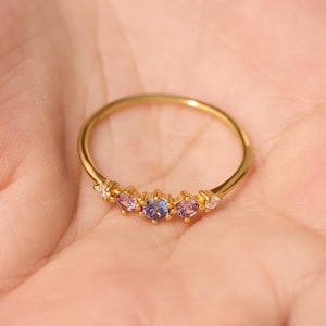 May include: A thin gold band ring featuring a row of five small, round-cut gemstones. The center stone is blue, flanked by two pink stones, and finished with two clear, white stones on the outer edges, all set in a delicate, polished gold metal setting.
