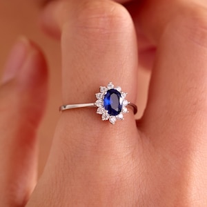 May include: A silver-toned ring featuring a central oval-cut deep blue gemstone surrounded by a halo of small, clear, round-cut stones. The band is thin and metallic, showcasing a classic cluster design that highlights the contrast between the blue centre and the surrounding clear stones.