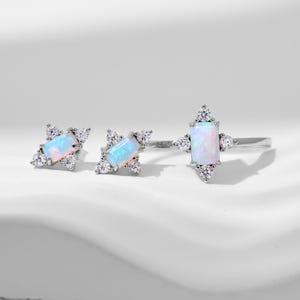 May include: A silver ring and two matching stud earrings, each featuring a rectangular iridescent opal center stone surrounded by four small clear round accent stones arranged in a star-like pattern on a polished silver metal band and setting.