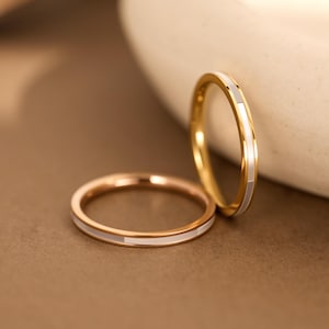 May include: Two thin gold rings featuring a smooth, polished finish and a continuous inlay of white rectangular segments. One ring lies flat while the other stands upright, showcasing the metallic band and the alternating white inlay pattern.