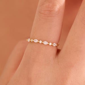 May include: A thin gold band ring featuring a delicate arrangement of alternating marquise-cut and round-cut clear stones set along the top half of the band, creating a continuous sparkling line across the finger.