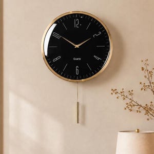 May include: A round wall clock with a black face, gold-tone frame, and gold hands. The clock features white numerals at the 12, 2, 4, 6, 8, and 10 positions, with thin white markers for other hours. A gold pendulum hangs from the bottom. The word Quartz is printed in white.