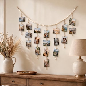May include: A wall-mounted photo display featuring a long string of small, light-colored wooden beads draped in a curve. Multiple vertical jute cords hang from the beads, each holding several small square photographs secured with tiny wooden clips.