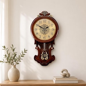May include: A wall clock with a dark brown wooden frame and a circular face featuring a gold-toned background, black Arabic numerals, and ornate central filigree. The lower section includes a decorative pendulum housing with gold-toned accents.