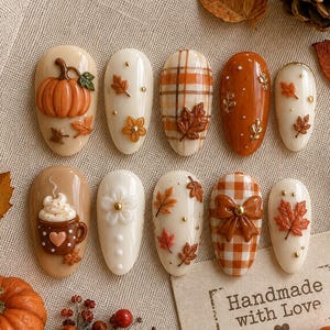 May include: A set of ten press-on nails featuring autumn-themed designs including a 3D orange pumpkin, brown plaid patterns, a mug of hot cocoa with whipped cream, orange and brown maple leaves, white flowers, and a brown gingham bow with gold beads.
