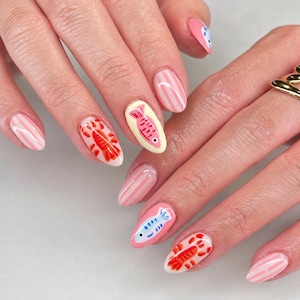 May include: Manicured fingernails featuring a mix of designs including light pink and white vertical stripes, orange lobster illustrations, and light blue or pink fish motifs on a pale yellow or pink base, with a gold ring on the right hand.