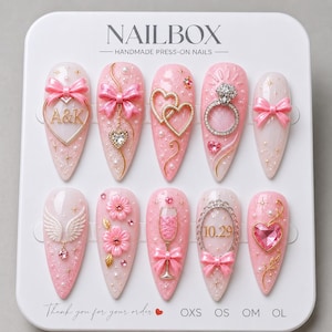 May include: A set of ten almond-shaped press-on nails on a white display card labeled NAILBOX. The nails are light pink with glitter, featuring 3D embellishments including pink bows, gold hearts with A&K, a diamond ring, wings, flowers, and a champagne glass.