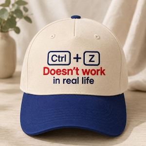 May include: A beige baseball cap with a dark blue brim and a top button. The front features embroidered text in dark blue and red that reads: Ctrl + Z Doesn't work in real life, with Ctrl and Z enclosed in dark blue rectangular outlines.
