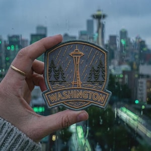 May include: A shield-shaped embroidered patch featuring a gray background with a gold-colored Space Needle, evergreen trees, and rain lines. The word WASHINGTON is stitched in gold capital letters along the bottom border of the patch.