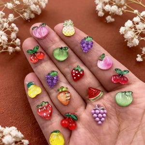 May include: A collection of small, colorful fruit and vegetable-shaped resin charms. The assortment includes miniature strawberries, grapes, cherries, apples, a pineapple, a pear, a carrot, a watermelon slice, and a lemon, all with glossy finishes.