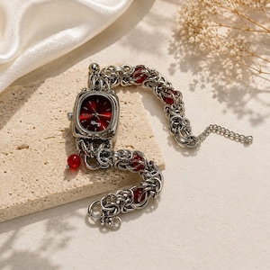 Puede incluir: Un reloj de pulsera plateado con una caja rectangular, esfera de color rojo intenso y el texto "Chaoyue Time". La correa está formada por complejos anillos metálicos entrelazados intercalados con pequeñas cuentas rojas translúcidas.