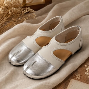 May include: A pair of split-toe shoes featuring a metallic silver front section and a cream-colored leather upper. The design includes side cutouts, a low-profile silhouette, and a thin silver trim along the collar, showcasing a distinct toe separation.