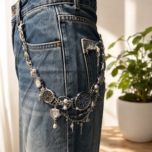 May include: A silver-toned metal chain accessory attached to blue denim jeans, featuring multiple integrated analog watch faces, small white pearl beads, and dangling charms shaped like seashells and small metallic objects.