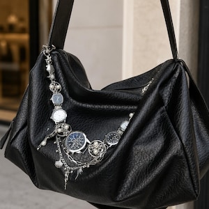May include: A black hobo bag with a textured surface and a long shoulder strap. The bag is adorned with a silver-toned chain accessory featuring multiple small analog wristwatches, white faux pearls, and small metallic seashell charms.