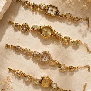 May include: Four gold-tone wristwatches with decorative link bracelets. The first has a square face, the second a round face with sun and shell charms, the third a heart-shaped face, and the fourth a round face with pearl-like accents.