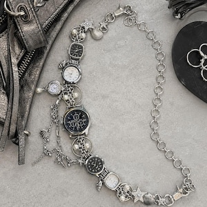 May include: A silver-toned chain necklace featuring multiple analog watch faces, small white pearls, and metallic star and seashell charms. The design incorporates various watch styles with black and white dials, connected by a series of circular links.