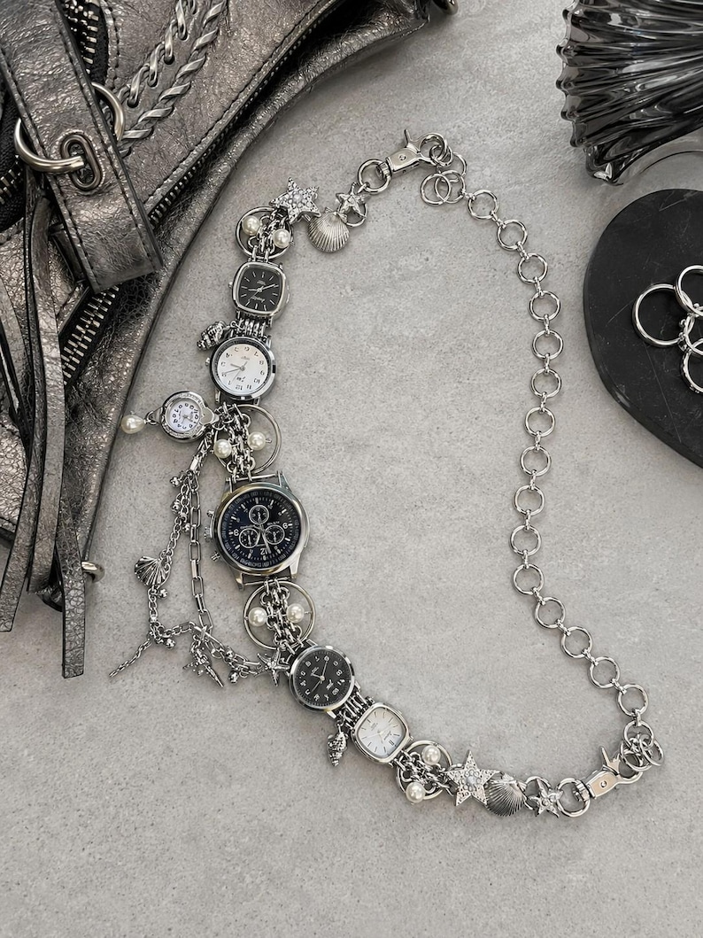 May include: A silver-toned chain necklace featuring multiple analog watch faces, small white pearls, and metallic star and seashell charms. The design incorporates various watch styles with black and white dials, connected by a series of circular links.