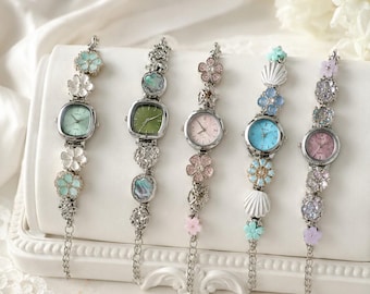 May include: Five silver-tone wristwatches with decorative floral and shell-shaped links. Each watch features a unique dial color including mint green, olive green, pale pink, bright blue, and soft pink, accented with crystals and metal flower charms.