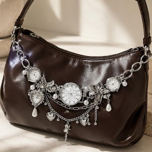 May include: A dark brown leather shoulder bag adorned with a silver-toned chain strap featuring three integrated analogue watch faces, dangling heart-shaped charms, faux pearl accents, crystal-encrusted floral elements, and a small rose pendant.