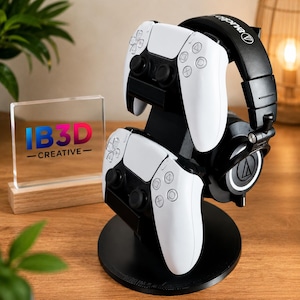 Pode incluir: Um suporte preto para acessórios de jogos com dois comandos brancos e um par de auscultadores pretos. Ao lado do suporte, encontra-se uma placa de acrílico transparente sobre uma base de madeira com o texto IB3D CREATIVE em azul, vermelho e preto.