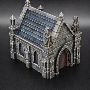 Peut inclure: Un bâtiment miniature en pierre avec un toit en bardeaux gris foncé, des fenêtres en arc et une porte en bois. La structure est ornée de deux silhouettes encapuchonnées sur les coins avant, avec une finition peinte en gris et marron.