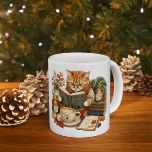 May include: A white ceramic mug featuring a graphic illustration of an orange tabby cat wearing glasses and a scarf while reading a book. The design includes a cup of coffee, a stack of books, a candle, and a notebook with autumn leaves.
