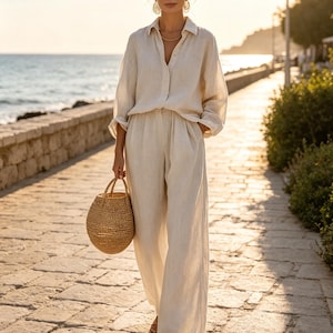May include: A beige long-sleeved button-down shirt and matching wide-leg trousers made of a textured fabric. The outfit is paired with a round woven straw handbag and simple flat sandals, creating a coordinated set in a neutral tone.