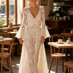 May include: A floor-length white lace dress featuring long bell sleeves, a deep V-neckline, and a fitted bodice that transitions into a flowing skirt. The intricate floral lace pattern covers the entire garment, creating a semi-sheer effect.