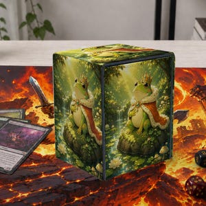 Puede incluir: Una caja para barajas rectangular con una rana verde que lleva una corona dorada y una capa roja, sentada sobre una roca cubierta de musgo. La caja descansa sobre un tapete con un paisaje de lava, cartas, un libro pequeño y varios dados.