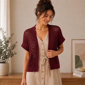 Può includere: Un cardigan all'uncinetto color bordeaux a maniche corte con un motivo a rete aperto e un bordo solido lungo le aperture frontali. Il capo è indossato sopra un abito beige chiaro abbottonato con dettaglio in vita e scollo a V.