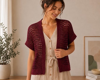 May include: A deep burgundy, short-sleeved crochet cardigan featuring an open-knit grid pattern and a solid border along the front edges. The garment is worn over a light beige, button-down dress with a tie-waist detail and a V-neckline.