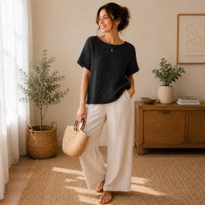 May include: A dark charcoal short-sleeved knit top with a round neckline paired with wide-leg beige trousers. The outfit is accessorized with a woven straw handbag featuring round wooden handles and simple brown flat sandals.