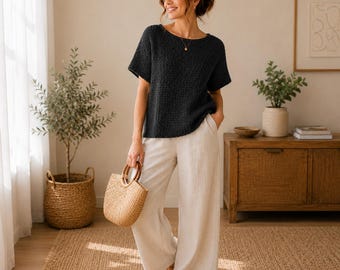 May include: A dark charcoal short-sleeved knit top with a round neckline paired with wide-leg beige trousers. The outfit is accessorized with a woven straw handbag featuring round wooden handles and simple brown flat sandals.