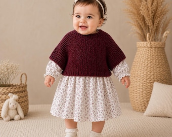 May include: A baby dress featuring a white floral print skirt with long sleeves and a dark burgundy knitted sweater overlay. The garment includes a round neckline and is paired with white shoes and a white headband with a small bow detail.