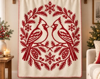May include: A cream-colored throw blanket featuring a red knit pattern of two cardinal birds facing each other. The design includes a circular wreath of pine branches, pinecones, berries, and snowflakes. The blanket has a red stitched border.