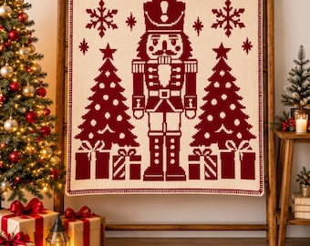 May include: A square knit blanket featuring a pixelated design in deep red on a cream background. The central motif is a nutcracker figure flanked by two Christmas trees and wrapped gift boxes, with snowflake accents above the trees.
