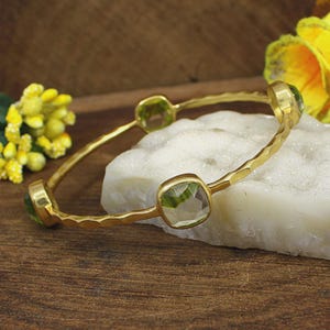 May include: A gold-toned bangle bracelet featuring a hammered metal texture and three square-cut, translucent green gemstones set at intervals along the band. The bracelet has a polished finish and a thin, flexible structure.